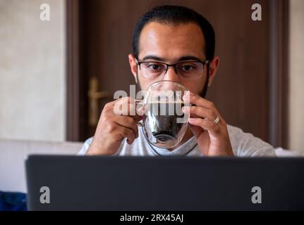 bearded male drinks coffee while working on his laptop from his home and eating cookies Stock Photo