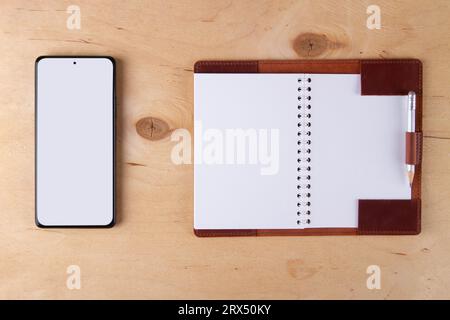 Digitalization concept. Paper notebook vs smartphone. Digital phone with blank white screen and empty jotter lie on wooden desk Stock Photo