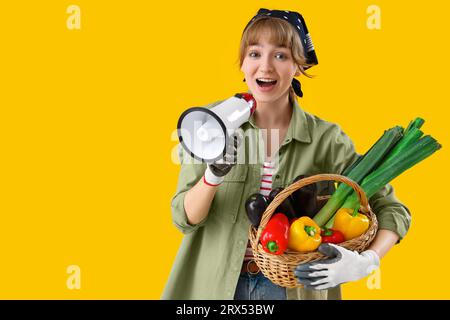 Young female farmer with megaphone and wicker basket of different ripe vegetables on yellow background Stock Photo
