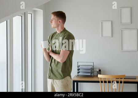 Pensive man with cup of coffee looking through apartment window Stock Photo