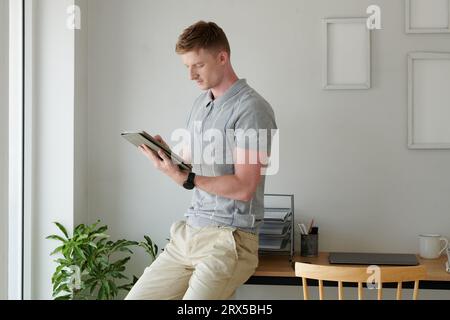 Young man sitting on desk and reading document or article on tablet computer Stock Photo