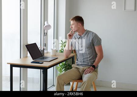 Pensive software developer sitting at office desk in his home office and looking through window Stock Photo
