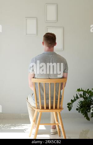 Young man sitting on chair and looking at empty picture frames hanging on wall Stock Photo