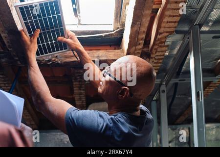 Construction worker examining solar panel over window in attic Stock Photo