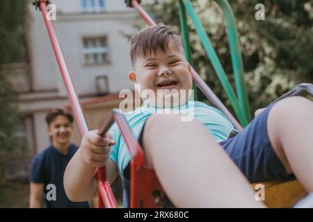 Happy boy with down syndrome playing on swing with brother in background Stock Photo