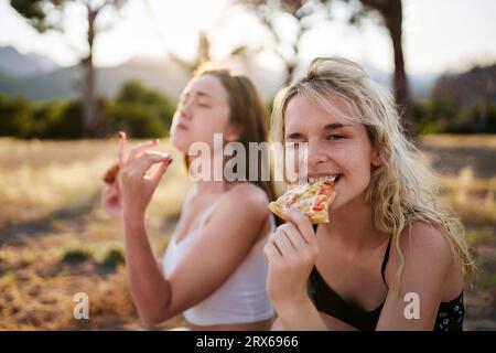 Young woman winking and eating pizza with friend in background Stock Photo