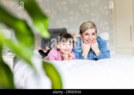 Smiling mother and daughter with hands on chins lying in bedroom Stock Photo