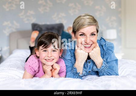 Happy mother and daughter with hands on chins lying in bedroom Stock Photo