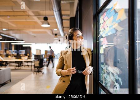 Businesswoman looking at adhesive notes on glass wall at office Stock Photo