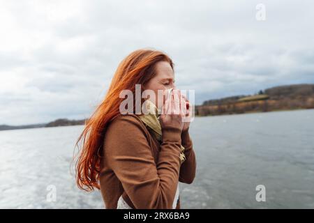 Redhead woman blowing nose by lake Stock Photo