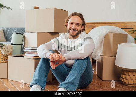 Happy young man sitting with hands clasped by cardboard boxes Stock Photo
