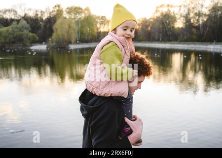 Brother carrying sister on shoulders by lake in park Stock Photo