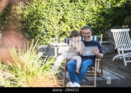 Happy man and boy using tablet PC sitting at back yard Stock Photo