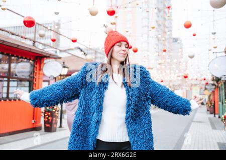 Smiling woman wearing Santa hat standing with arms outstretched on street Stock Photo