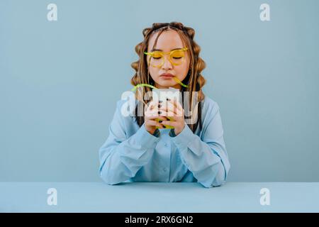 Young woman drinking cocktail through glass-like straw against blue background Stock Photo