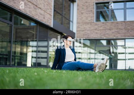 Businesswoman with VR headset sitting on grass at office park Stock Photo