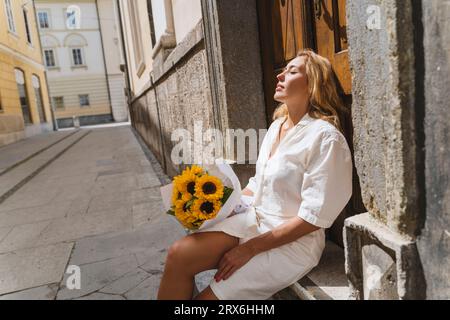 Blond woman sitting with eyes closed and sunflowers on sunny day Stock Photo
