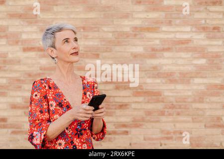 Contemplative woman with smart phone in front of wall Stock Photo