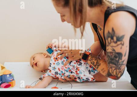Young, tattooed mother putting her baby on a bed at medical office, getting it ready for medical check up. Little baby girl playing with toys before c Stock Photo