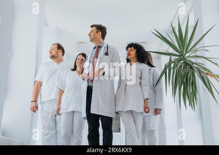 Group of confident doctors in medical uniforms walking through a hospital hall, looking out the window. Stock Photo