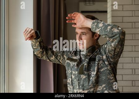 Serious short-haired young man in military uniform soldier looking through the window, upset soldier suffering from posttraumatic syndrome after Stock Photo
