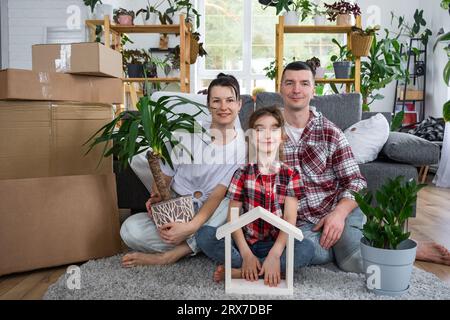 Moved family with a child and home plant are enjoying a new home, sitting on the sofa and holding the roof near boxes with things. Mortgage, insurance Stock Photo