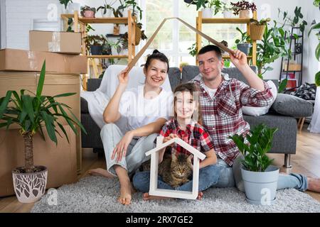 Moved family with a child and a cat pet are enjoying a new home, sitting on the sofa and holding the roof near boxes with things. Mortgage, insurance, Stock Photo