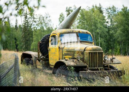 Old military equipment. Abandoned rusty yellow heavy war truck with big missile on board stands near the fence in an overgrown field and green trees at the background. Stock Photo