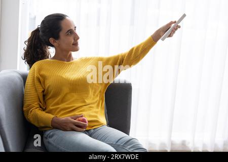 Hipanic indian woman using air conditioner remote control change adjust room temperature for comfortable home living room in summer season Stock Photo