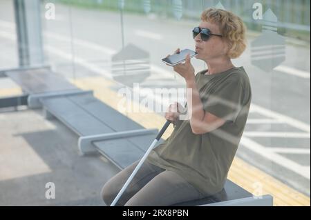 Elderly blind woman sitting at a bus stop and using a smartphone.  Stock Photo