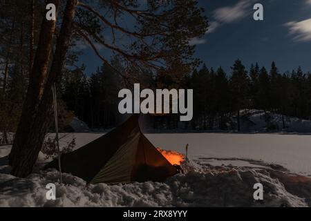 Traditional shelter with a campfire in front set up on the ice near the shore of a small snowy lake in winter in Finland Stock Photo