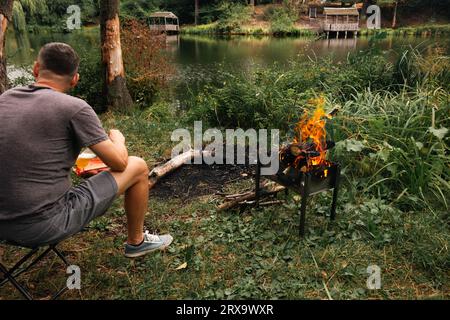 Man sitting in forest near bonfire. Man on a fall picnic. Lonely man on riverbank with campfire. Tourism in woods. Tourist near fireplace on pond bank Stock Photo
