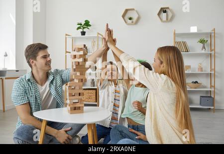 Happy family with two children are playing tumbling tower giving high five sitting on floor at home. Stock Photo