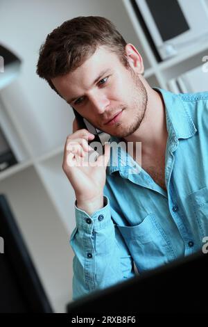 A young handsome man is talking on a mobile phone. A Caucasian guy is sitting in a room and talking on the phone. Vertical portrait. Stock Photo
