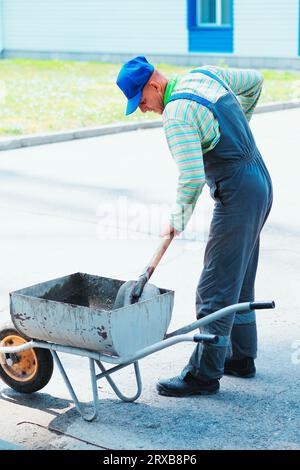 Male builder mixes cement mortar with shovel outside on summer day. Worker works at construction site. Builder prepares cement. Stock Photo