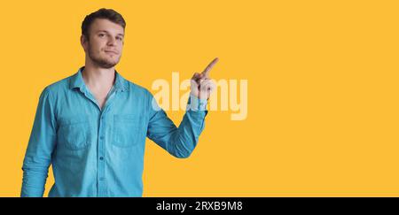 Man points his finger at empty space on yellow background. Portrait of handsome young Caucasian guy in denim shirt isolated on yellow background. Layout for inserting information. Stock Photo