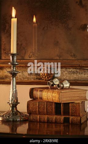Antique leather-bound books next to a silver candelabra In the background is an antique Venetian mirror with antique opera glasses resting on books. Stock Photo