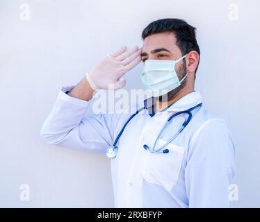 Doctor doing military salute on isolated white background holding his stethoscope wearing mask during healthcare day Stock Photo