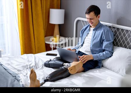 positive male with disability and special needs sit on bed and work on laptop. cheerful and excited. Handicapped Caucasian male using laptop for work Stock Photo