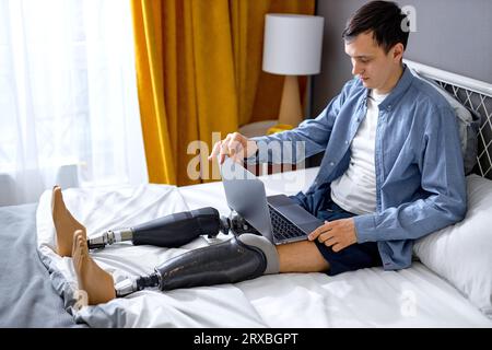Young worker with disability and special needs sit on bed opening laptop for job. Serious and concentrated. Handicapped Caucasian male using laptop fo Stock Photo