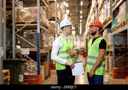 warehouse managers in work wear organizing distribution in warehouse storage area. Caucasian men in green uniform discuss product delivery, having tal Stock Photo