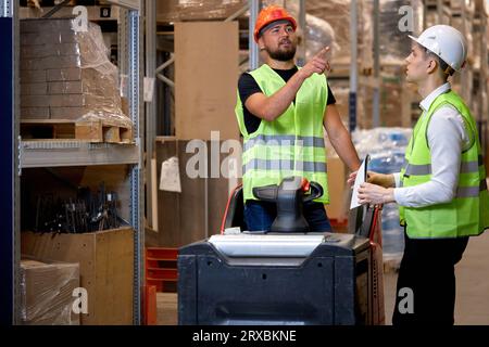 Young caucasian men workers doing stocktaking of products on shelves in warehouse together, using clipboard and forklift, having talk discussing, cont Stock Photo