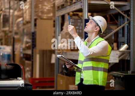 logistic business, shipment and people concept. male in helmet with clipboard checking goods at warehouse, counting and controlling distribution. conf Stock Photo
