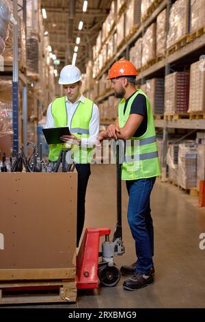 caucasian men warehouse workers with hand pallet jack lift stand having talk, temwork. hand pallet truck and male mover. Workers in uniform with cargo Stock Photo