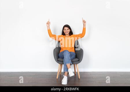 Positive young eastern woman in armchair pointing up, copy space Stock Photo