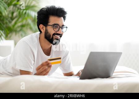 Online Payments. Smiling Indian Man Using Laptop And Credit Card In Bed Stock Photo