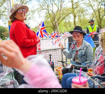 Two women smiling & celebrate whilst camping on the Mall London UK ahead of the coronation of King Charles dressed in Union Jack glasses waving flags Stock Photo