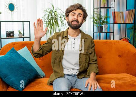 Hello. Middle eastern handsome man smiling friendly at camera and waving hands gesturing hello hi, greeting or goodbye, welcoming with hospitable expression at home. Guy sitting on sofa in living room Stock Photo
