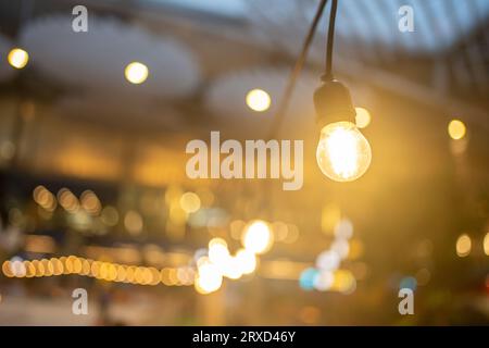 Decorative outdoor light bulbs hanging in air. Cozy warm yellow light bulbs lined up in row, background is blurred. Luminous incandescent lamps hanging in form go garland in yard.  Stock Photo