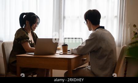 Two happy young Asian college students are working remotely at a coffee shop together, using laptop, doing their homework or co-project, preparing for Stock Photo